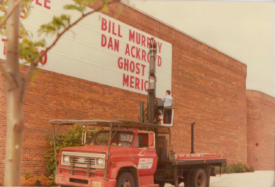 A red truck with a lift holding a man painting a sign advertising Ghostbusters with Bill Murray and Dan Ackroyd.