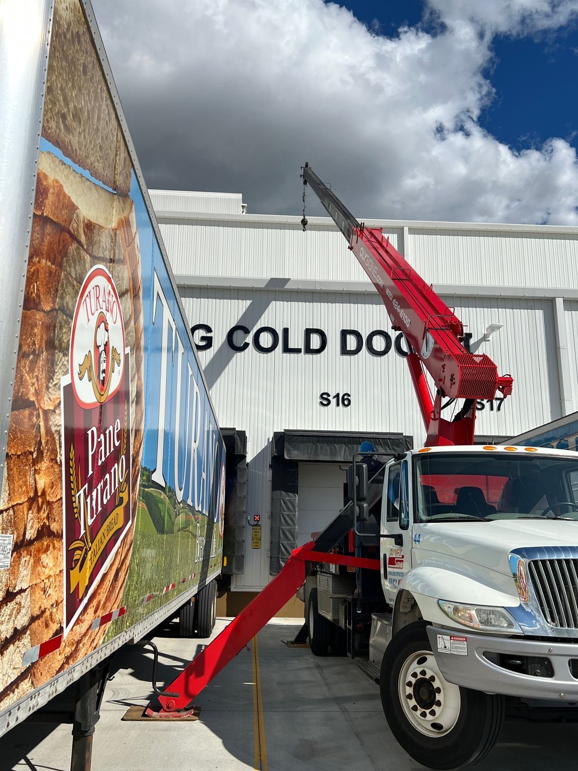 A red crane truck is parked next to a refrigerated trailer at a loading dock labeled