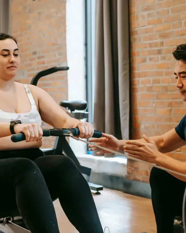 A man is helping a woman exercise on a rowing machine in a gym.