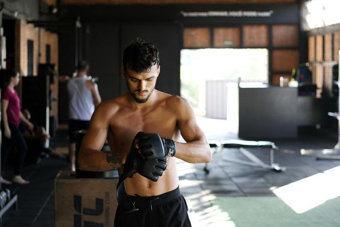 A shirtless man is wrapping his hands in boxing gloves in a gym.
