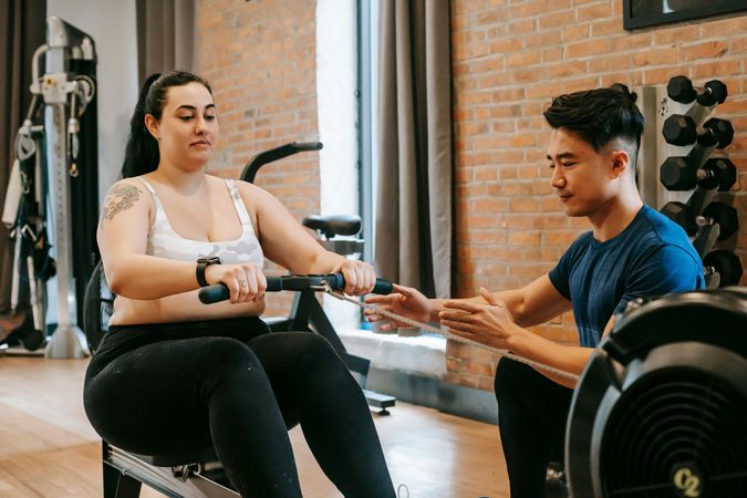 A man is helping a woman exercise on a rowing machine in a gym.