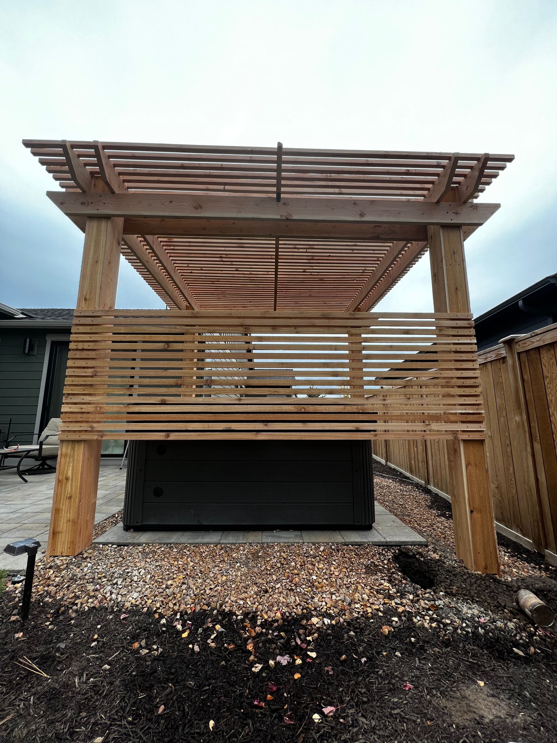A wooden pergola with a hot tub underneath it in a backyard.