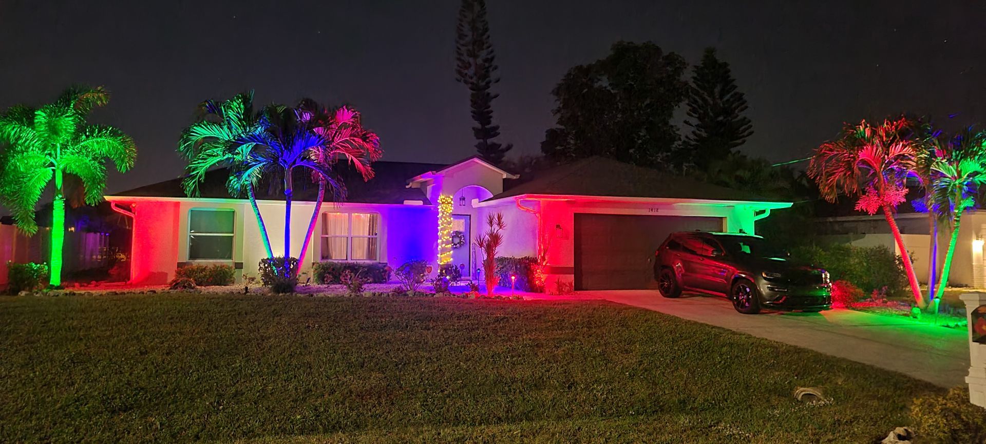 House with colorful lights illuminating palm trees and a car in the driveway at night.
