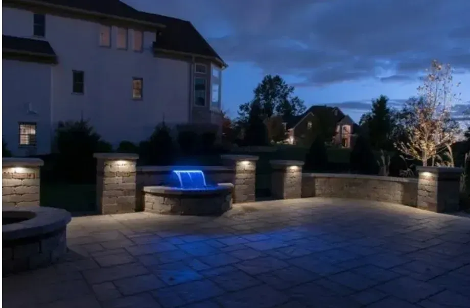 Night scene: Lit stone patio with a waterfall feature and a large house in the background.