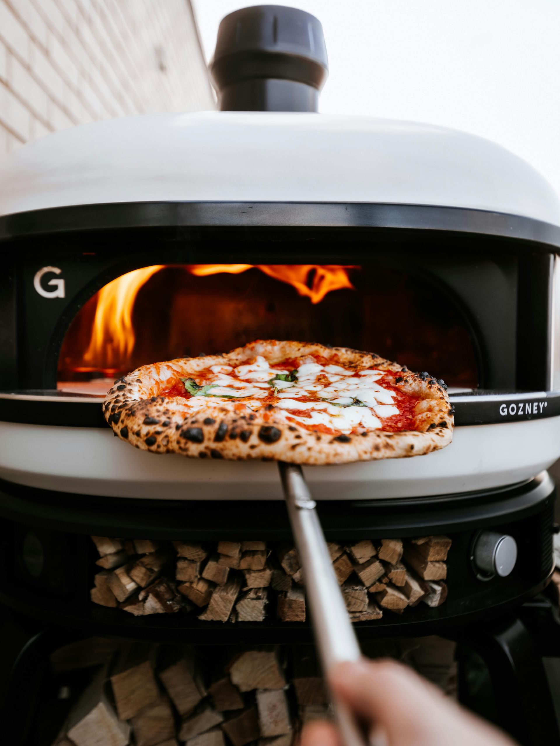 Pizza being pulled from a white outdoor pizza oven with a roaring fire, held by a metal pizza peel.
