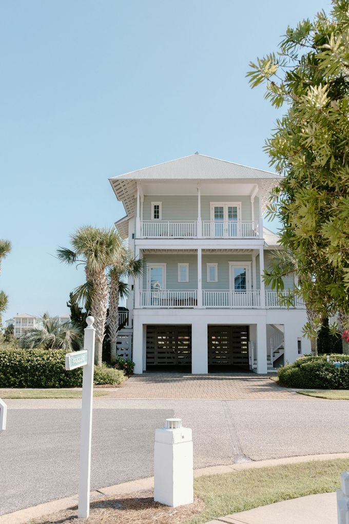 Two-story light green beach house with white trim, gravel driveway, and a blue sky.