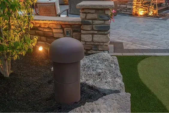 Brown trash can next to stone wall with dark mulch. Green grass and patio in background.