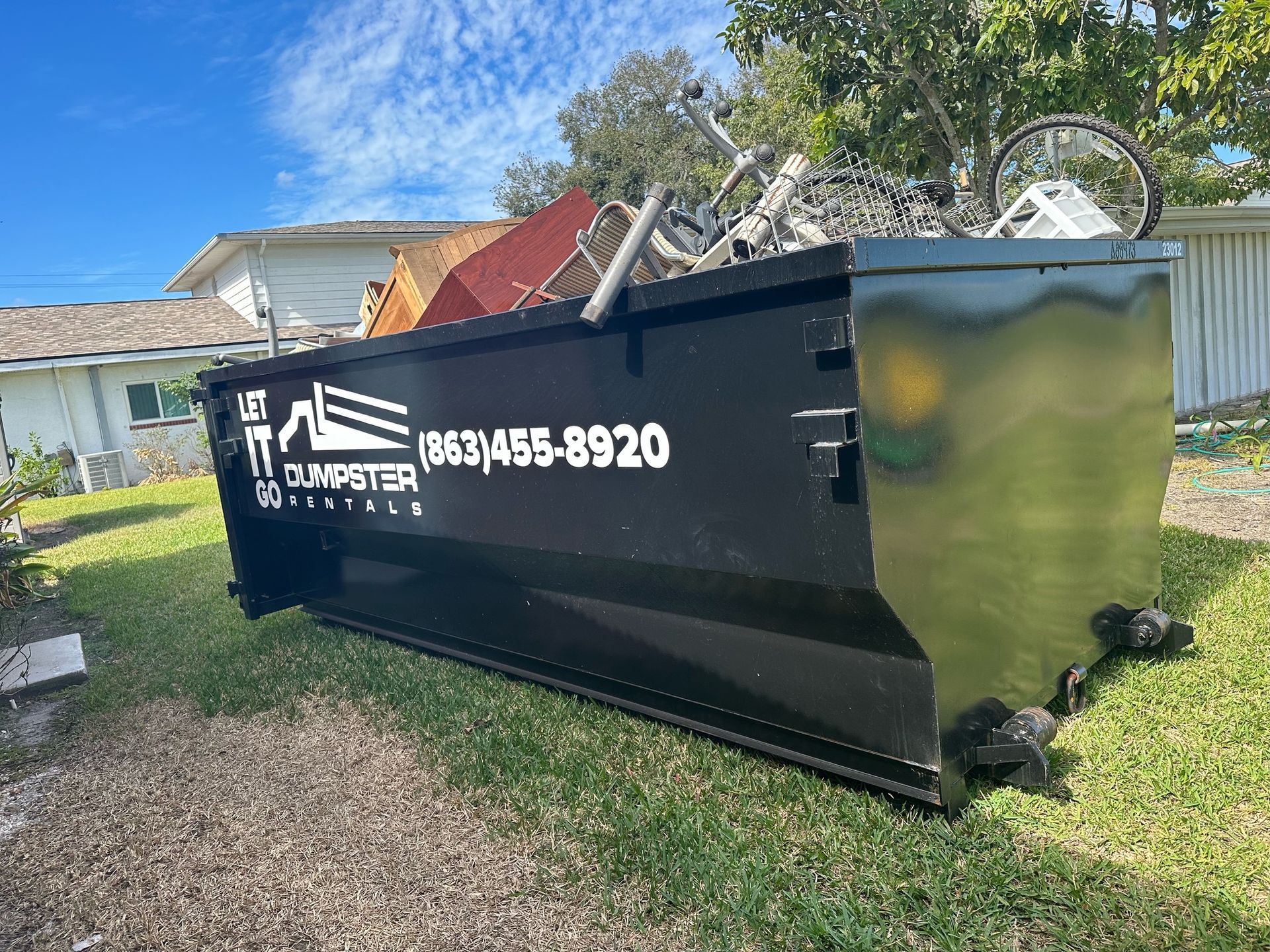 A large black dumpster is sitting in the grass in front of a house.
