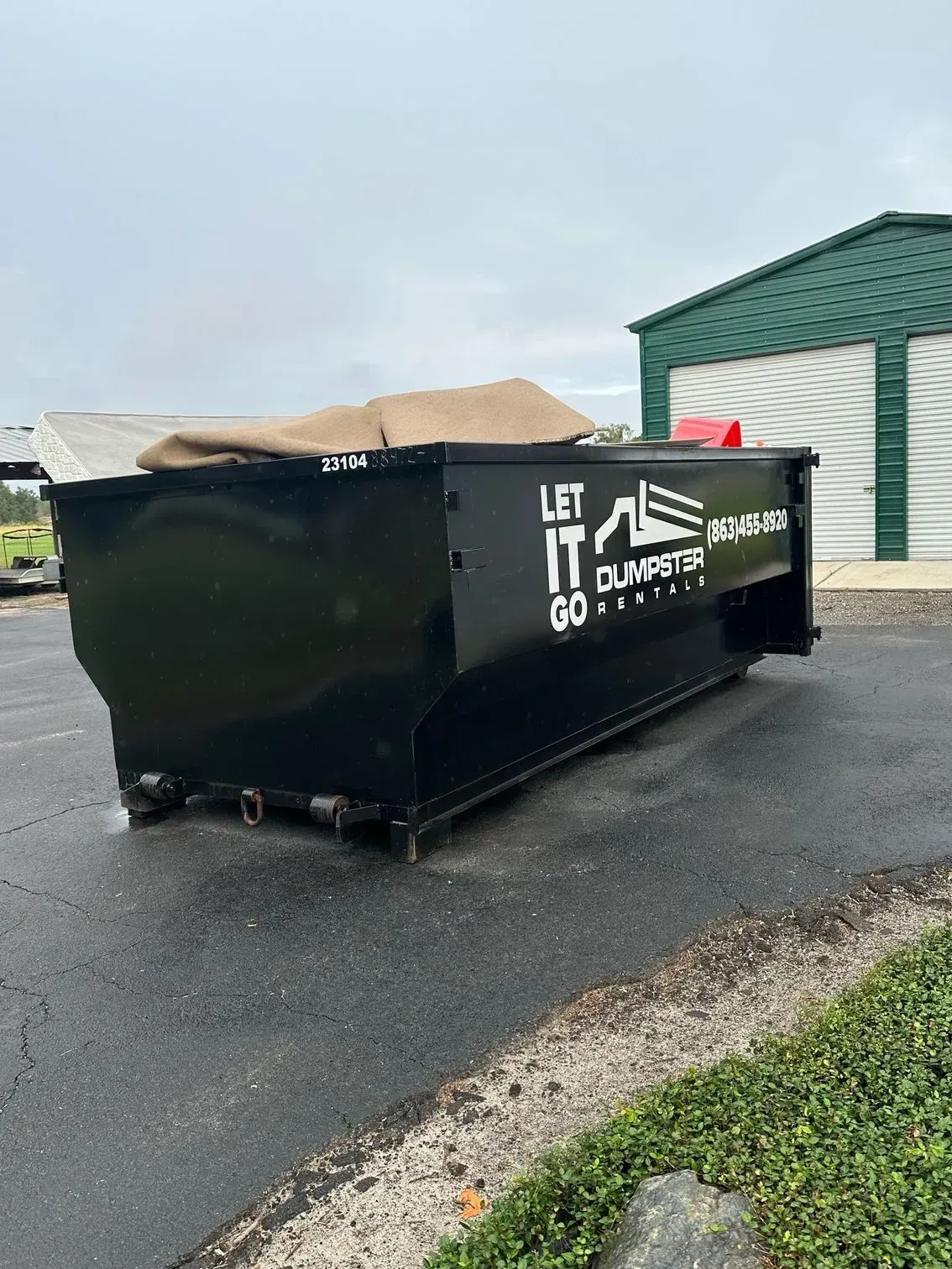 Black dumpster with open cardboard lid beside a green building on wet pavement