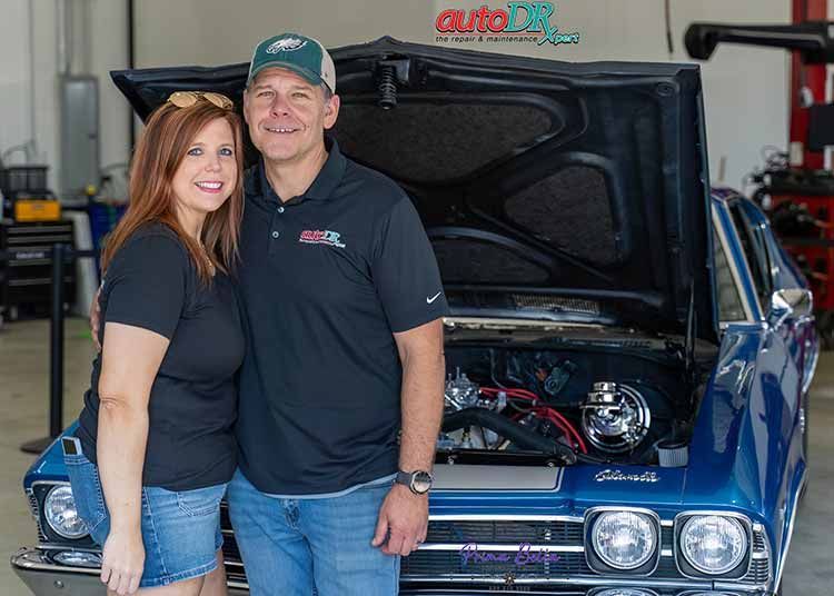 The owners of Auto DR smile while standing in front of a vintage car.