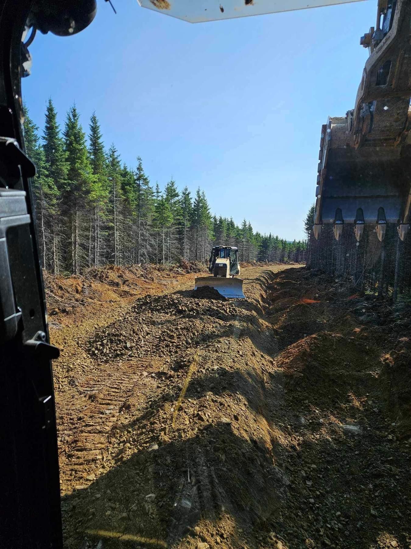 Excavatrice dégageant un chemin à travers une forêt, bulldozer au loin, ciel bleu.
