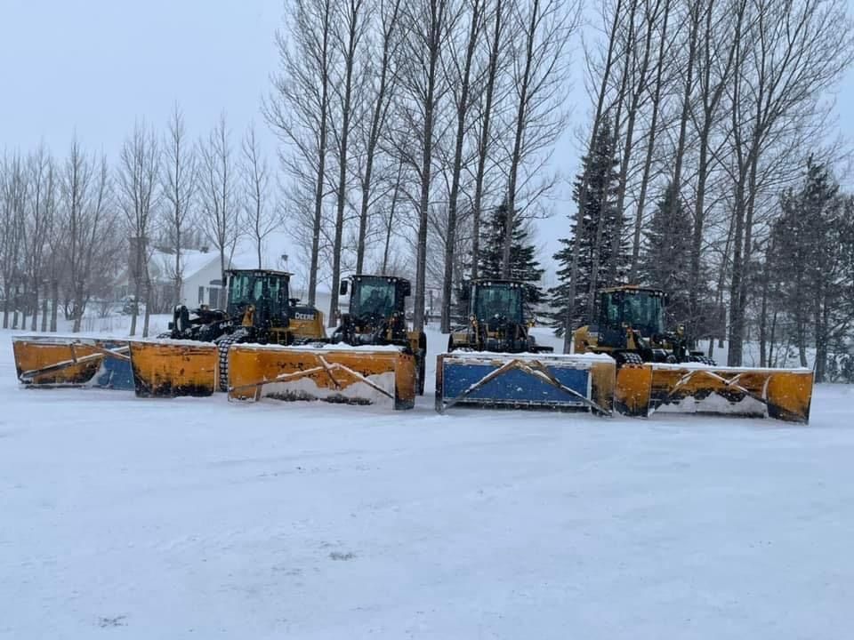 Quatre souffleuses à neige jaunes alignées, prêtes à déblayer la neige devant des arbres nus et un bâtiment.