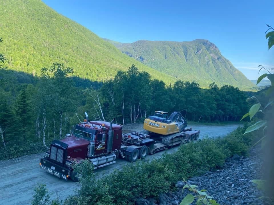 Semi-camion rouge transportant une excavatrice jaune sur une remorque, avec en arrière-plan les montagnes.