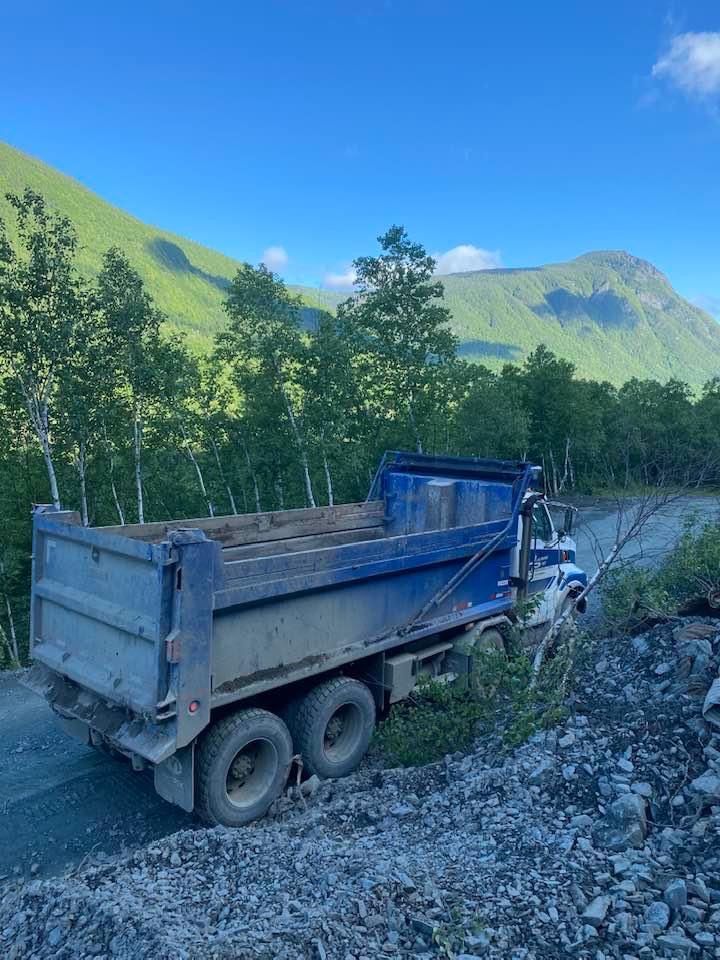 Camion à benne basculante bleu sur une route de gravier, montagnes et arbres en arrière-plan sous un ciel bleu.