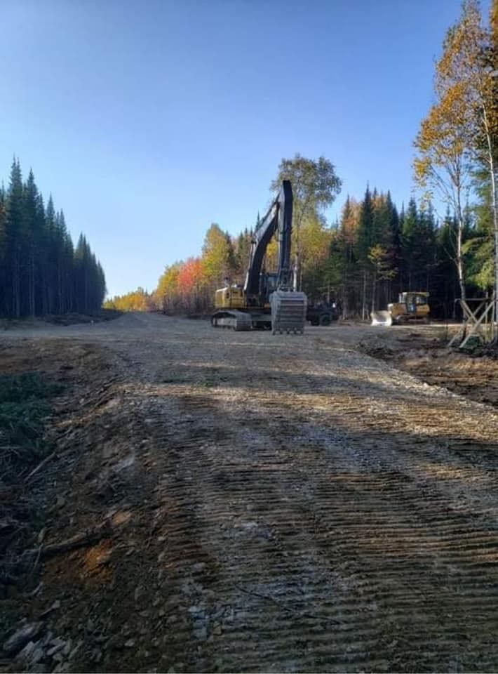 Un chemin de terre en construction à travers une forêt, avec une pelleteuse au centre. Ciel bleu.