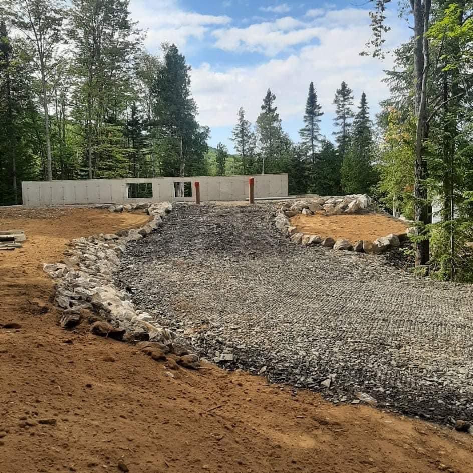 Allée de gravier menant à une fondation en béton dans une zone boisée par une journée ensoleillée.