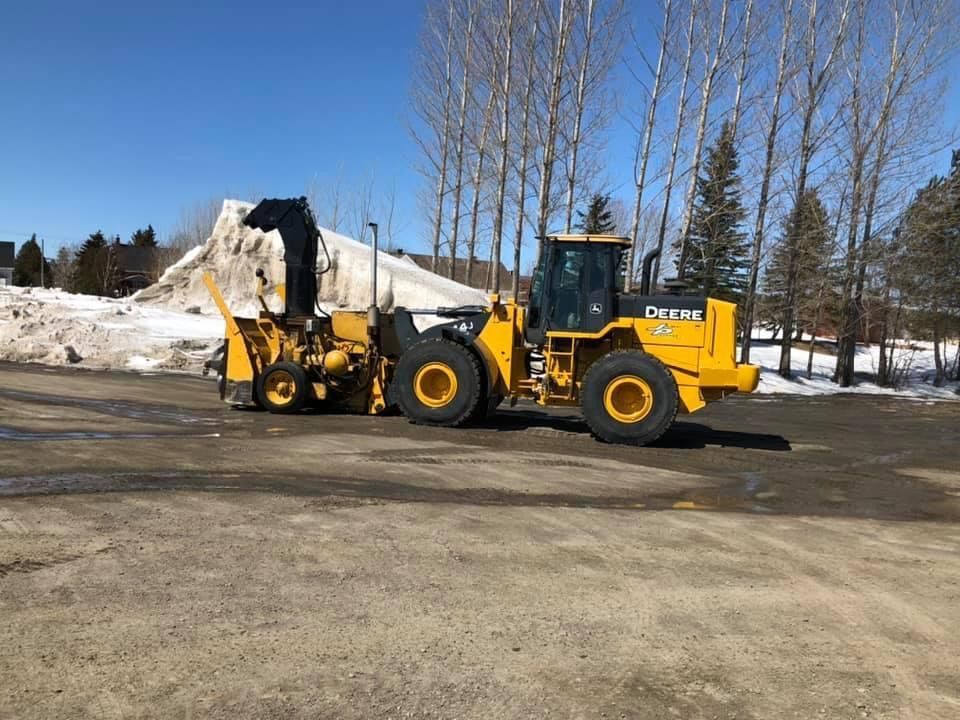 Chargeur jaune John Deere avec souffleuse à neige déneigeant une zone pavée, arbres en arrière-plan.