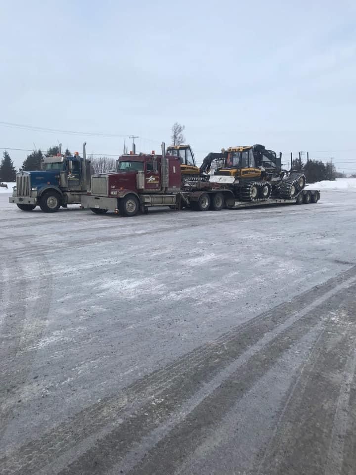 Deux semi-remorques stationnés sur une surface enneigée; l'un rouge avec une remorque transportant de la machinerie lourde.