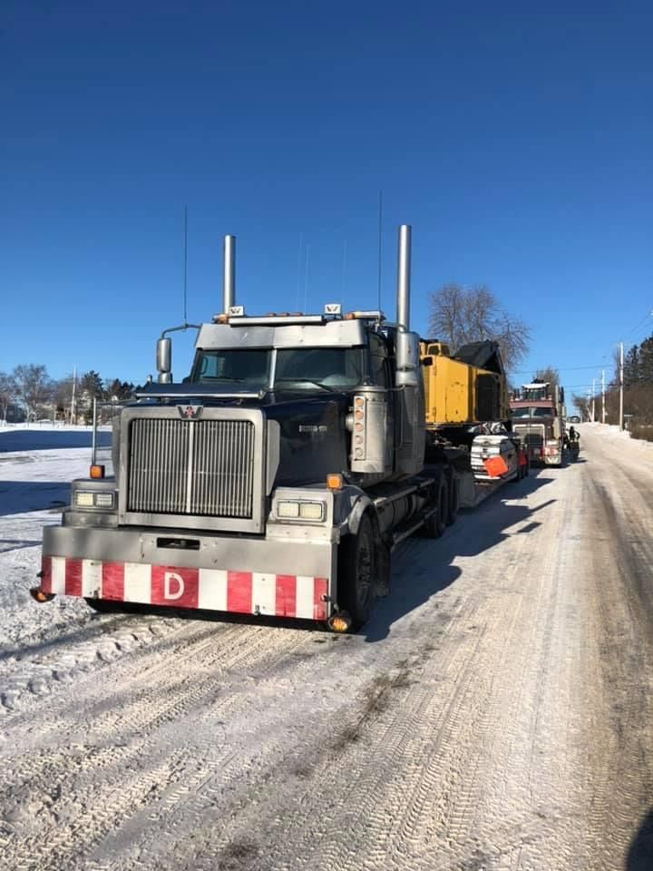 Semi-camion noir et argent avec une remorque jaune, roulant sur une route enneigée par une journée ensoleillée.