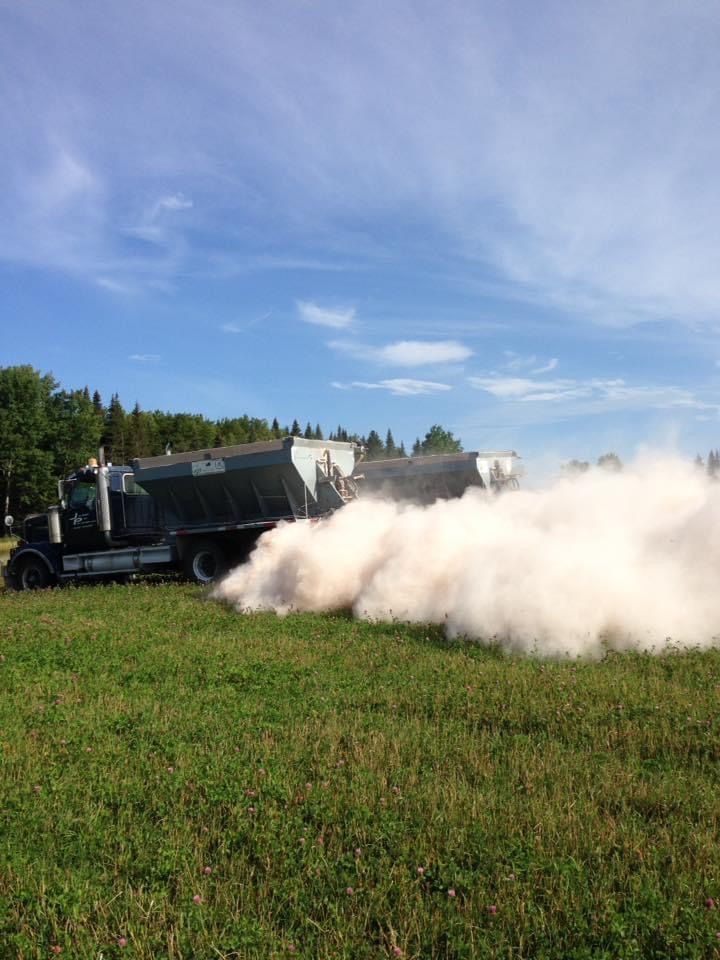 Un camion répand un nuage de poussière blanche sur un champ vert sous un ciel bleu.