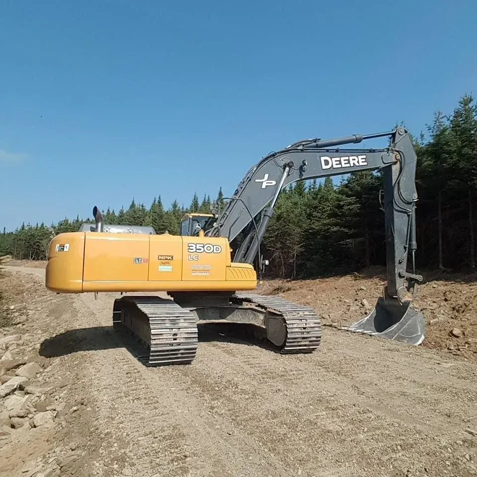 Excavatrice John Deere jaune sur un chemin de terre avec des arbres et un ciel bleu.