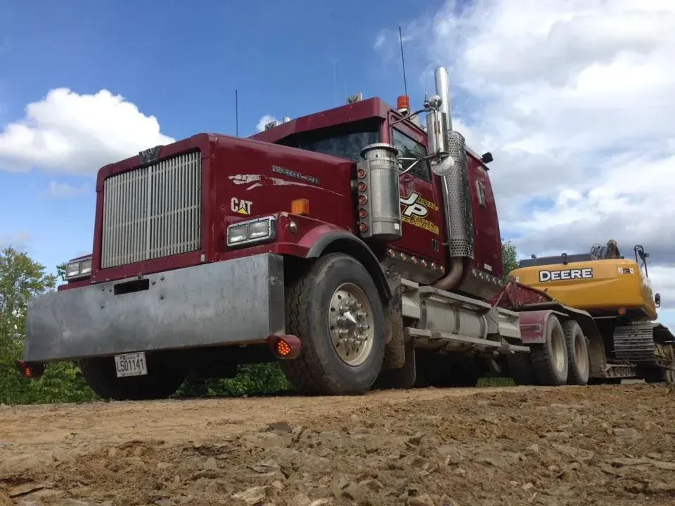 Semi-camion rouge transportant une excavatrice jaune sur un chemin de terre sous un ciel bleu.