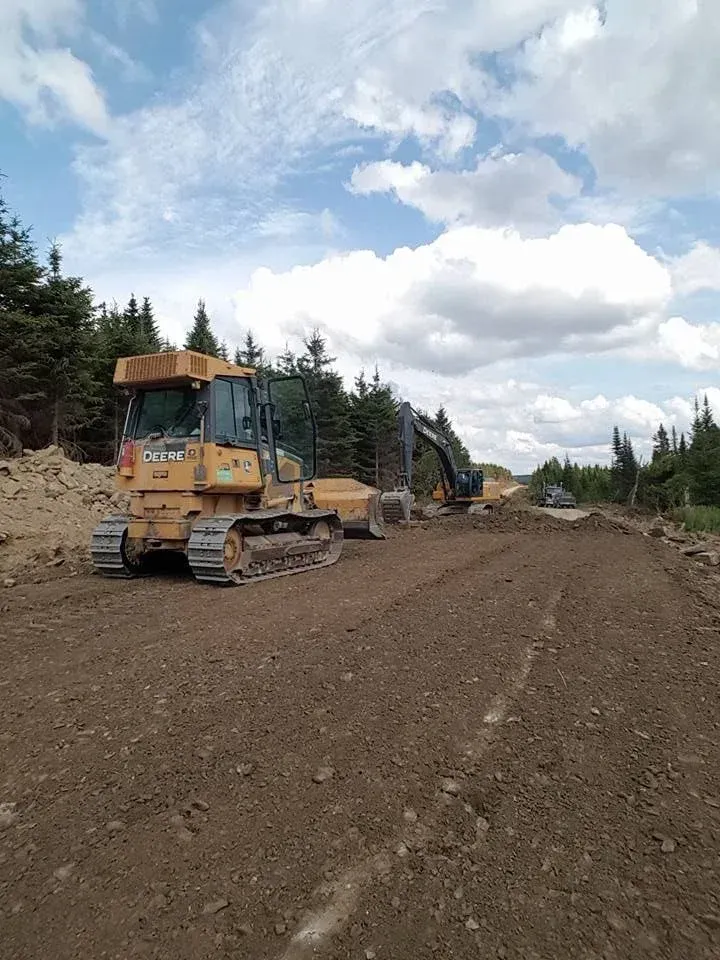 Un bulldozer et une excavatrice sur un chemin de terre, défrichant un terrain sous un ciel nuageux.