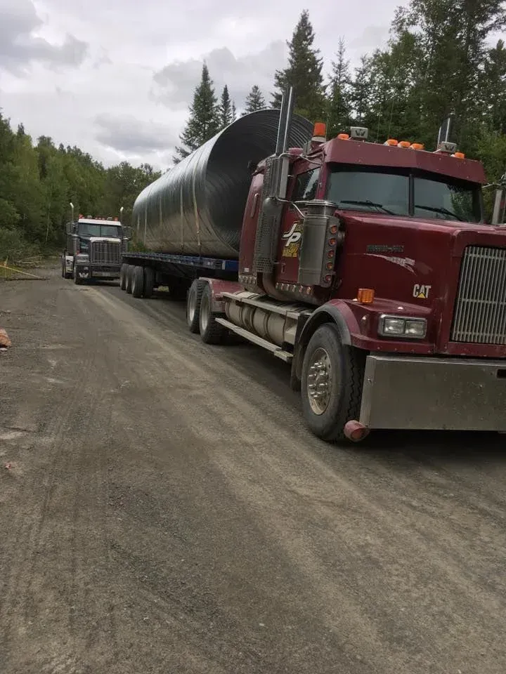 Semi-camion rouge transportant un réservoir cylindrique argenté sur une route de gravier, un autre camion en arrière-plan.