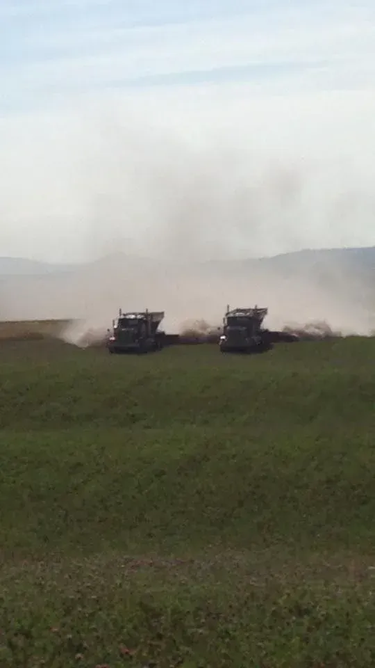 Deux camions épandant de l'engrais sur un champ, soulevant de la poussière. Paysage vallonné en arrière-plan.