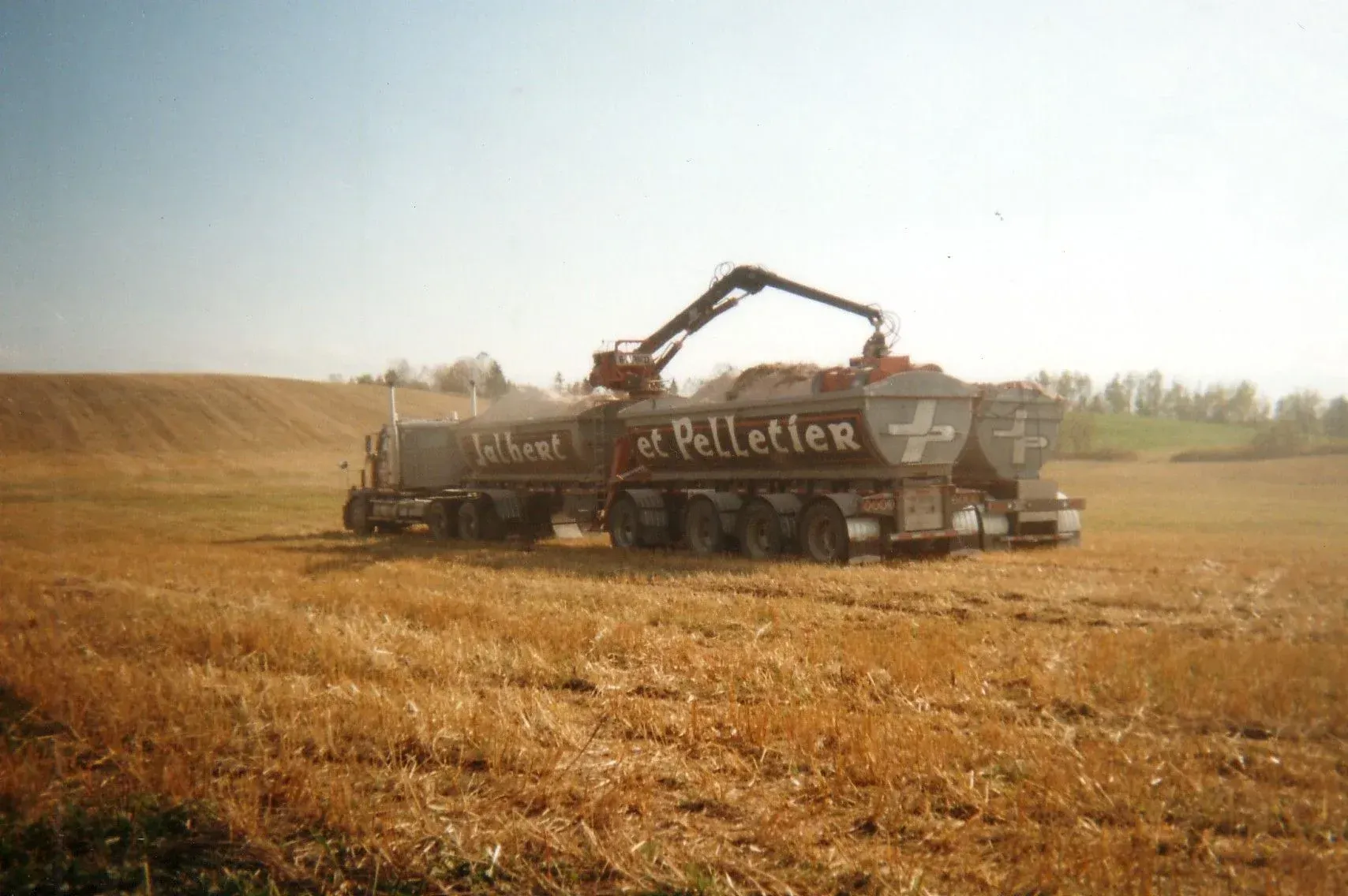 Camion chargeant du blé récolté dans un champ sous un ciel bleu; le camion porte le nom « Rheault et Pelletier ».