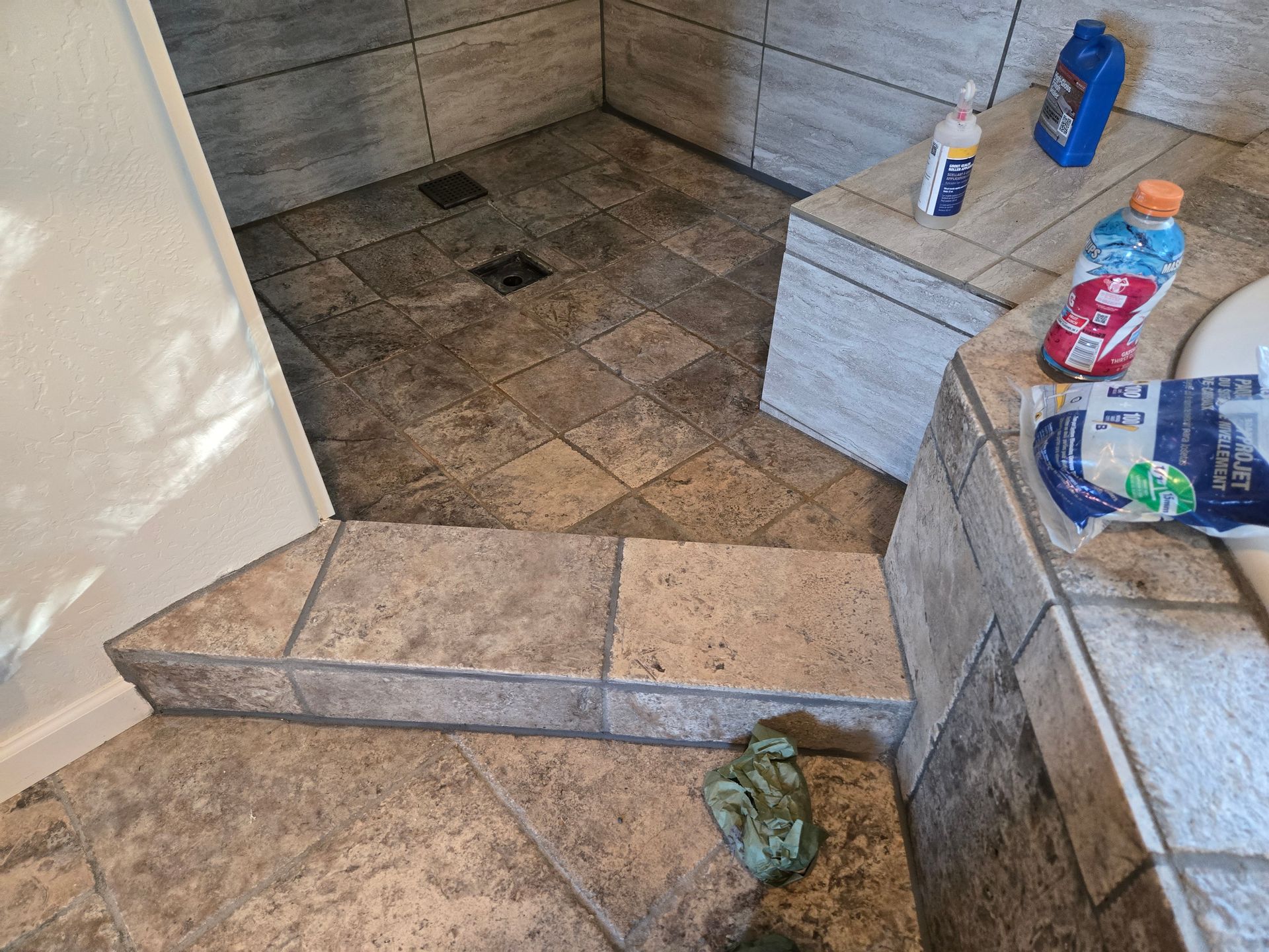 Small tiled shower with a low step, wooden bench, and toiletries on the ledge.