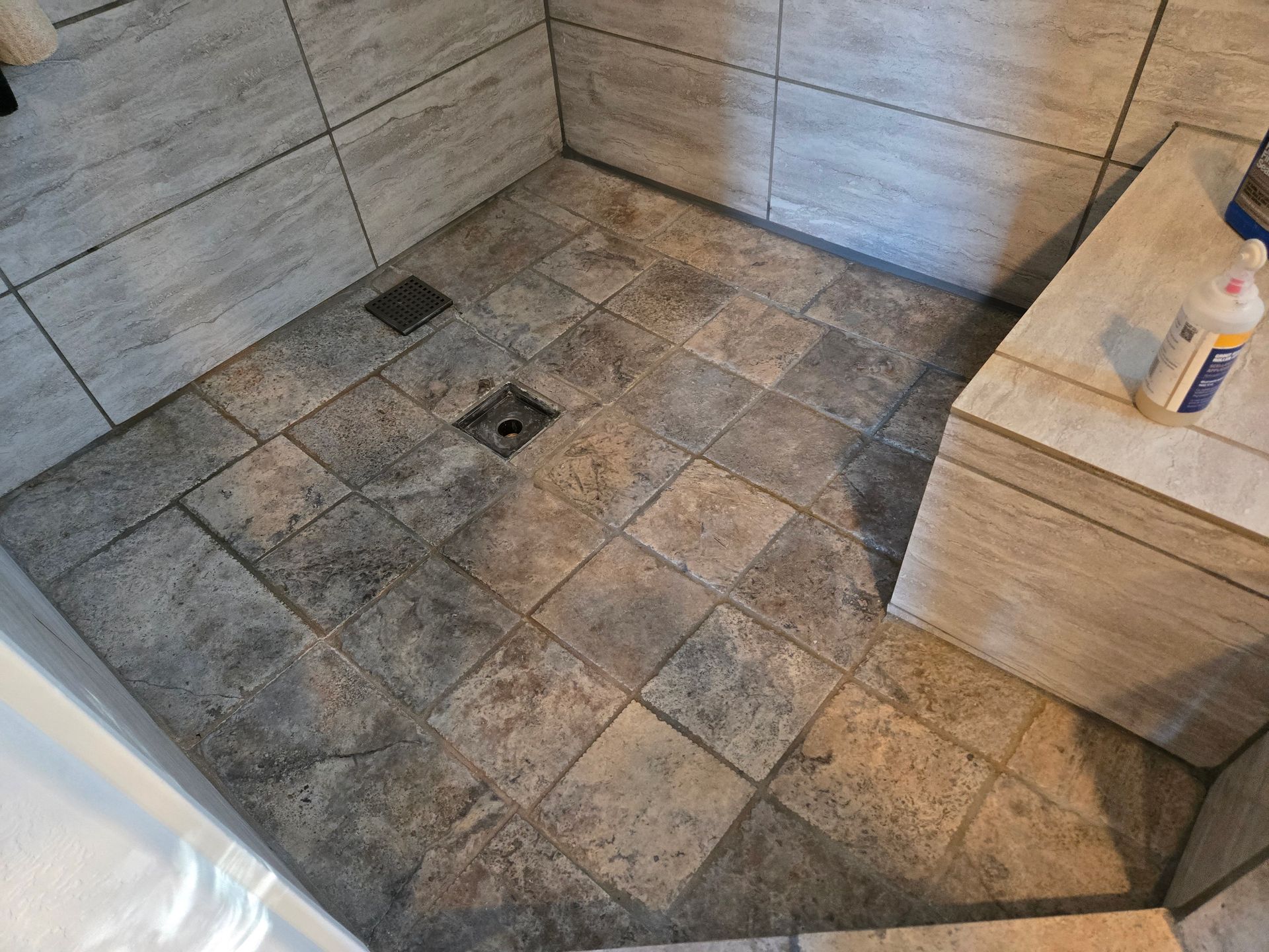 Tiled shower floor with a square drain and a wooden bench holding cleaning supplies.