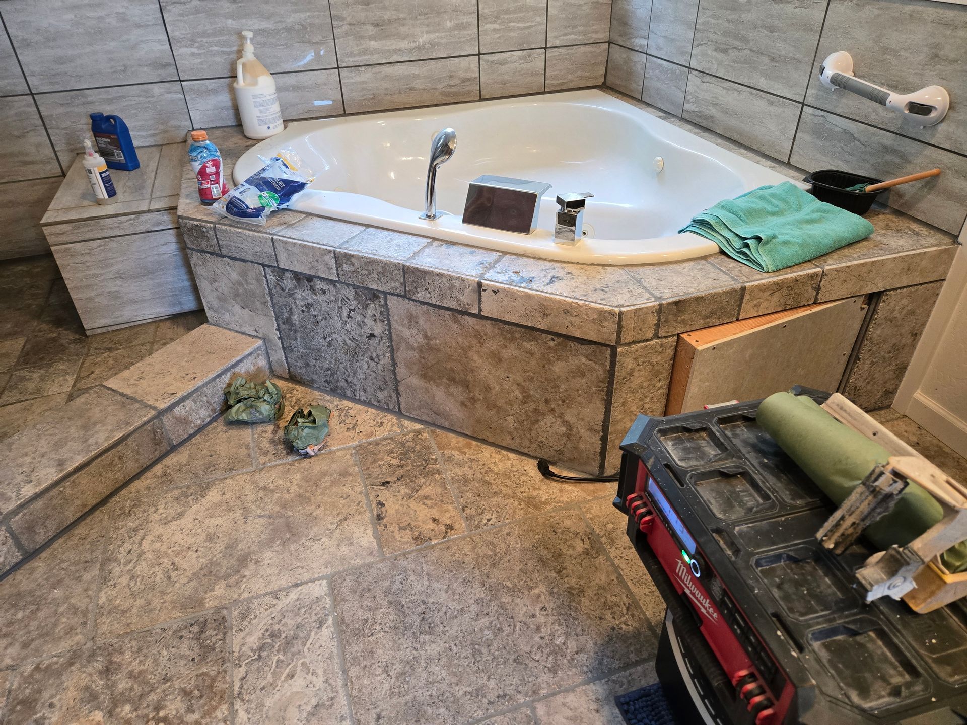 Bathroom with a large corner jacuzzi tub, stone tile walls and floor, and cleaning supplies nearby