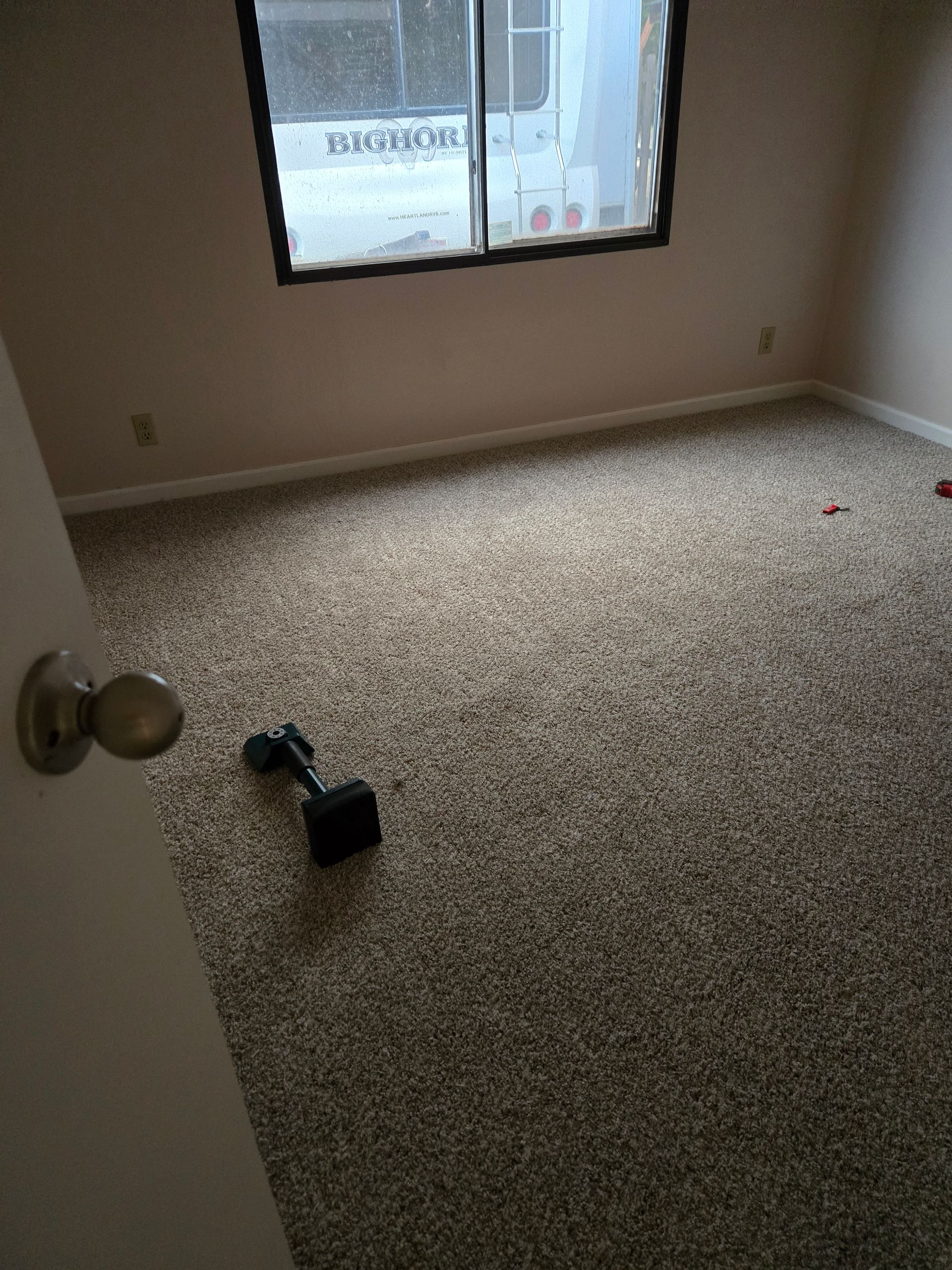 Empty beige room with carpet, window, and a doorknob in the foreground; a small item lies on the floor.
