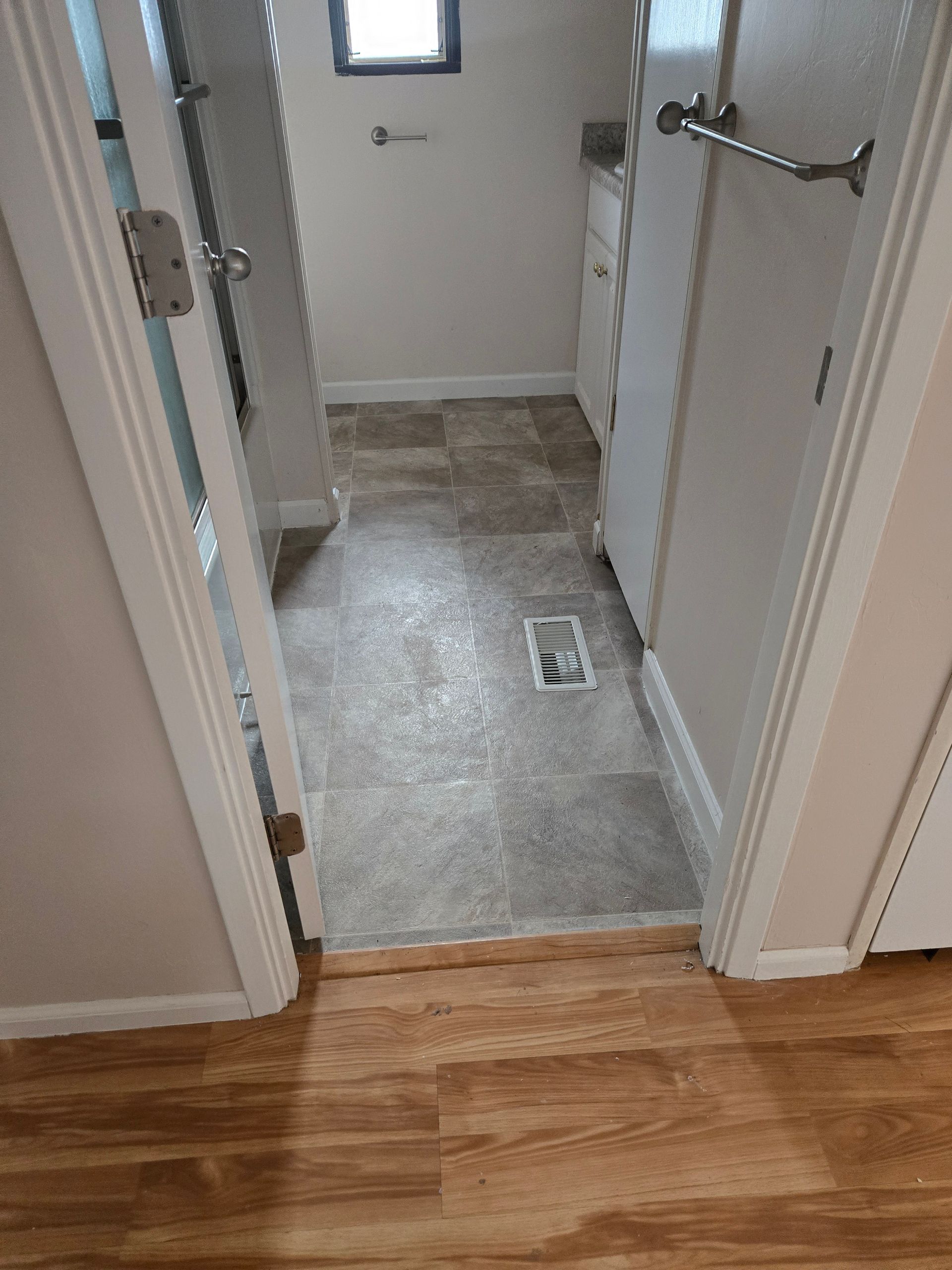 Hallway view into a bathroom with gray tile floor, white walls, and wood flooring in the foreground.