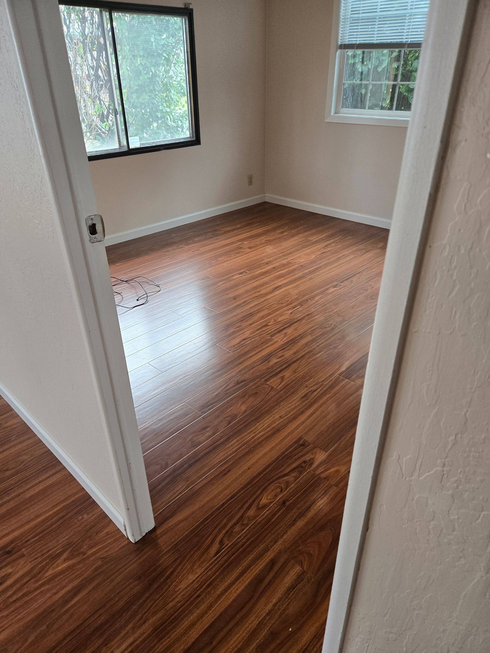 Empty room with wood floors and two windows, viewed through an open doorway.