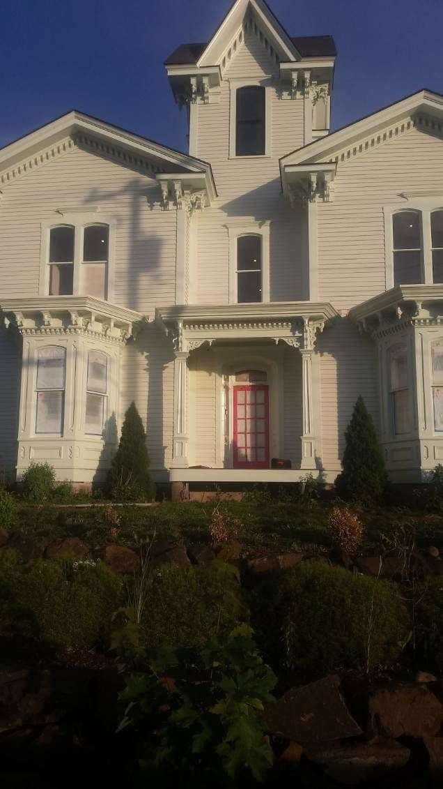 White Victorian-style house with tall gables, a front porch, and a red front door, partly shaded by trees.