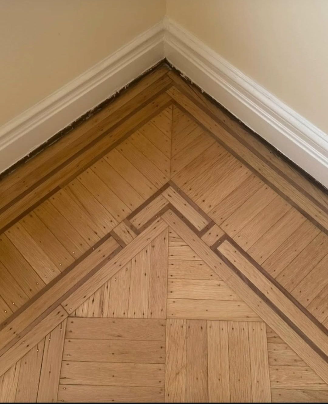 Wood parquet floor corner with decorative border pattern beside white baseboards and beige walls