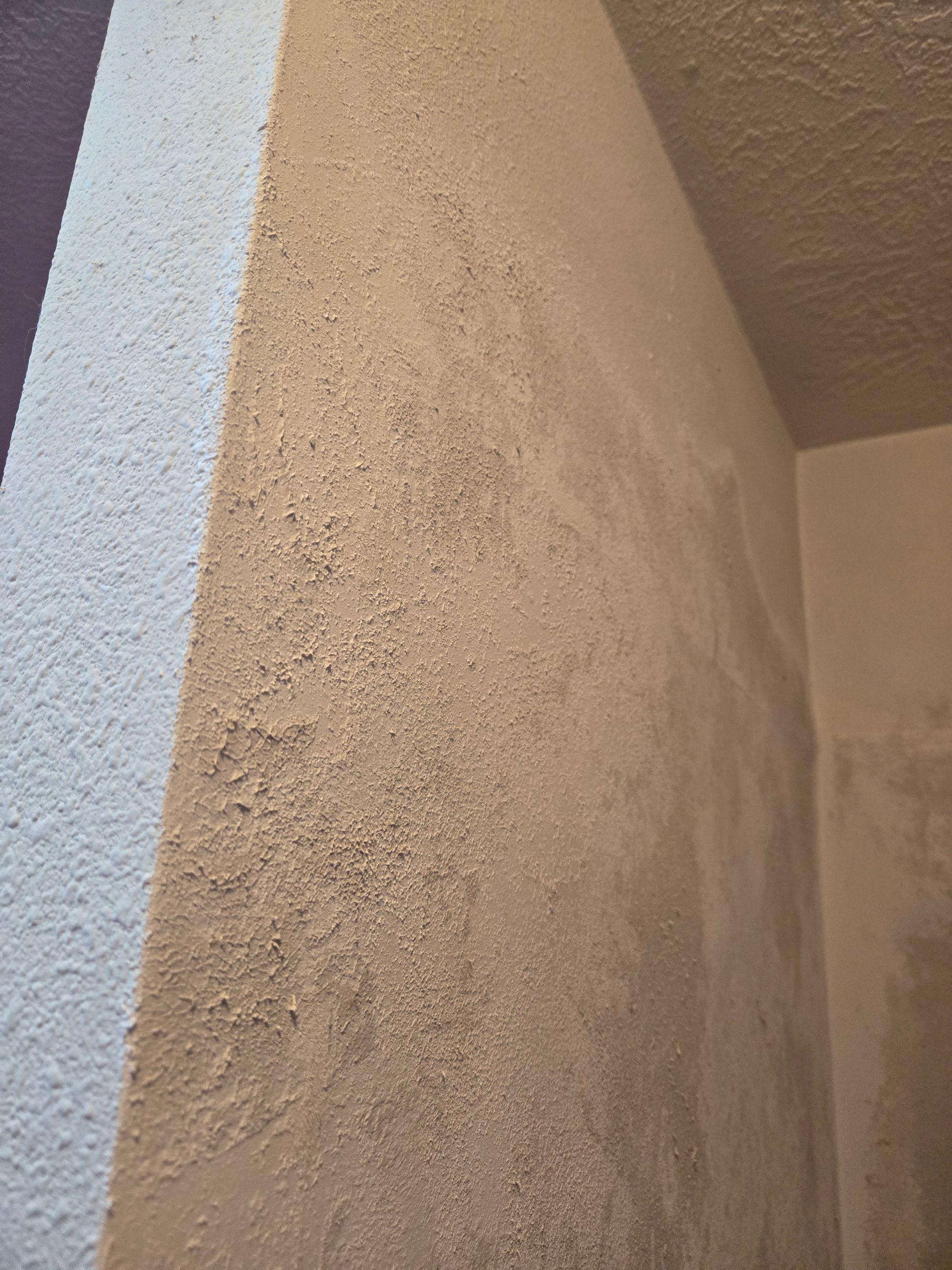 Close-up of a beige stucco wall corner beside a light gray wall and ceiling.