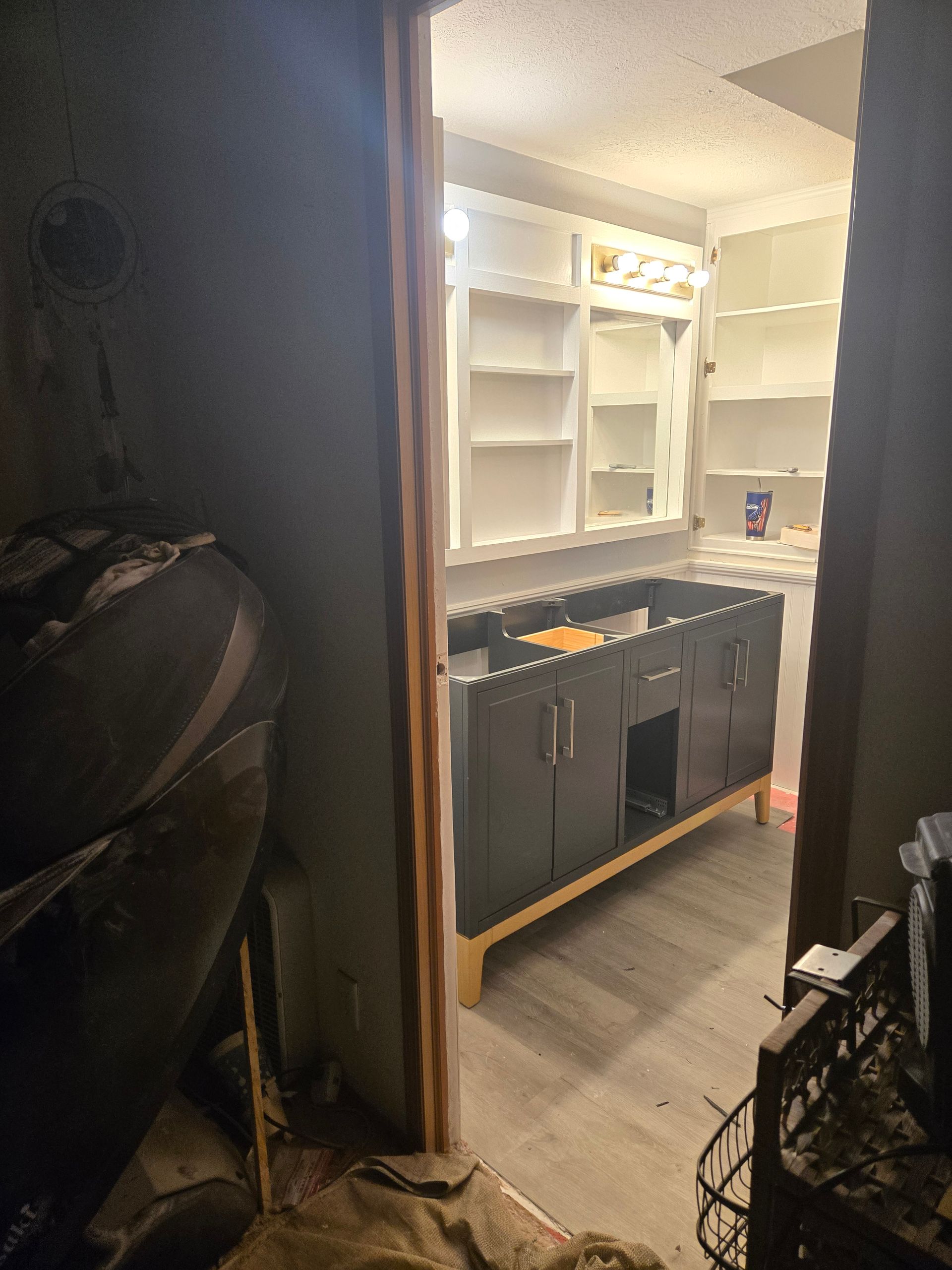 Bathroom with dark vanity, white shelves, and mirror lit by bright lights, viewed through a doorway