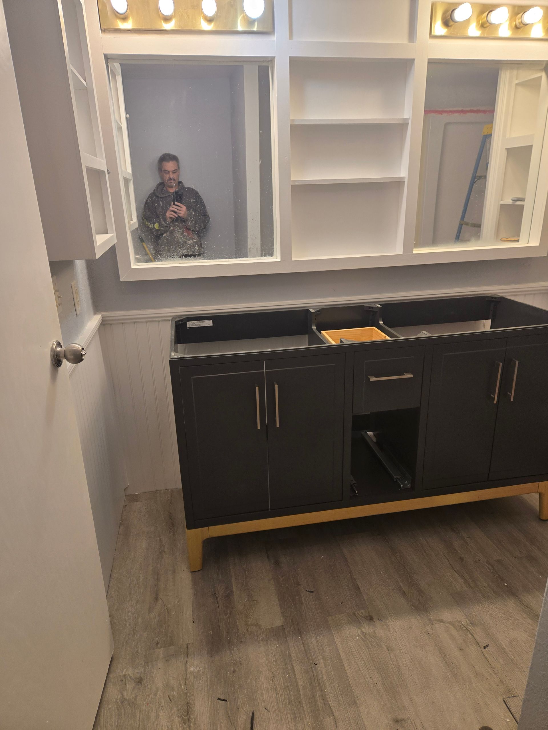 Bathroom vanity with dark cabinets, white shelves, and a mirror reflecting a person holding the camera.