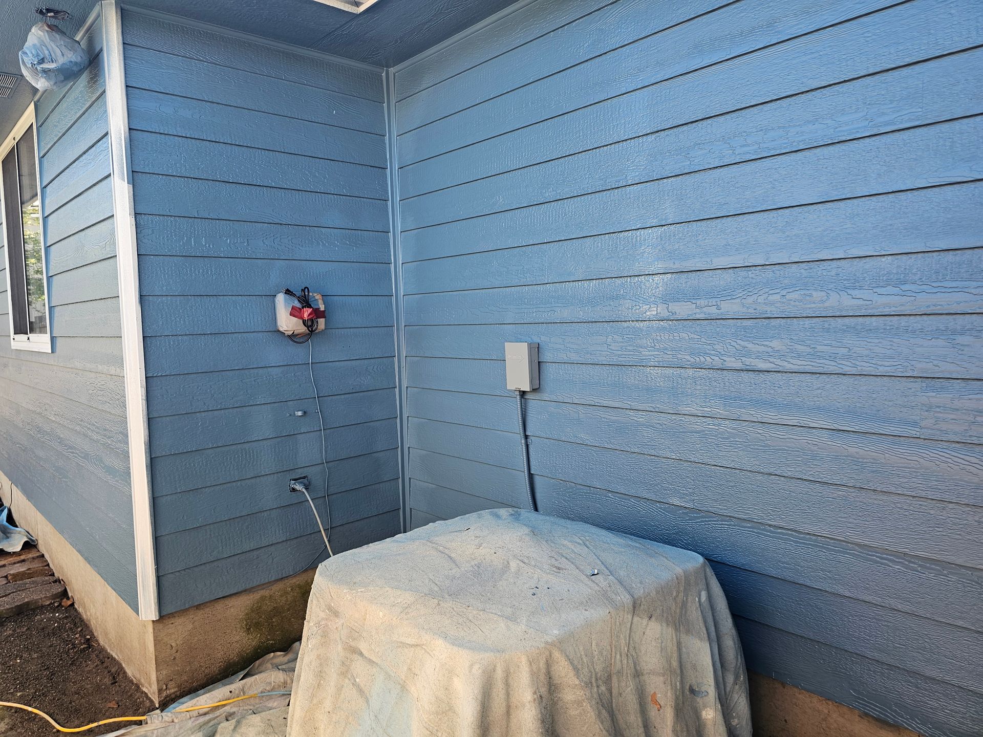 Blue house exterior corner with a covered appliance on a small patio and a wall outlet nearby