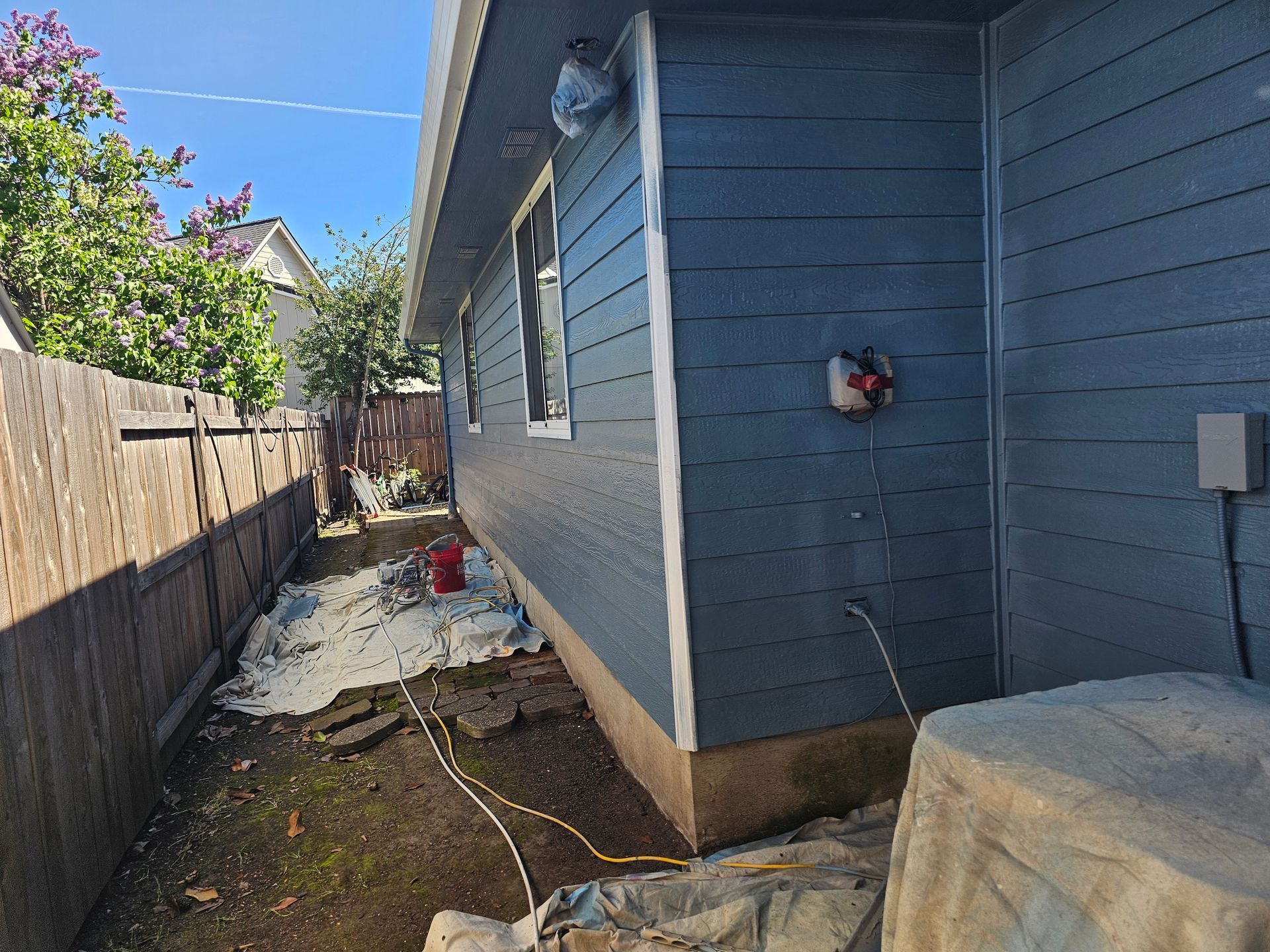 Narrow side yard beside a blue house with white tarp or debris piled along the fence.