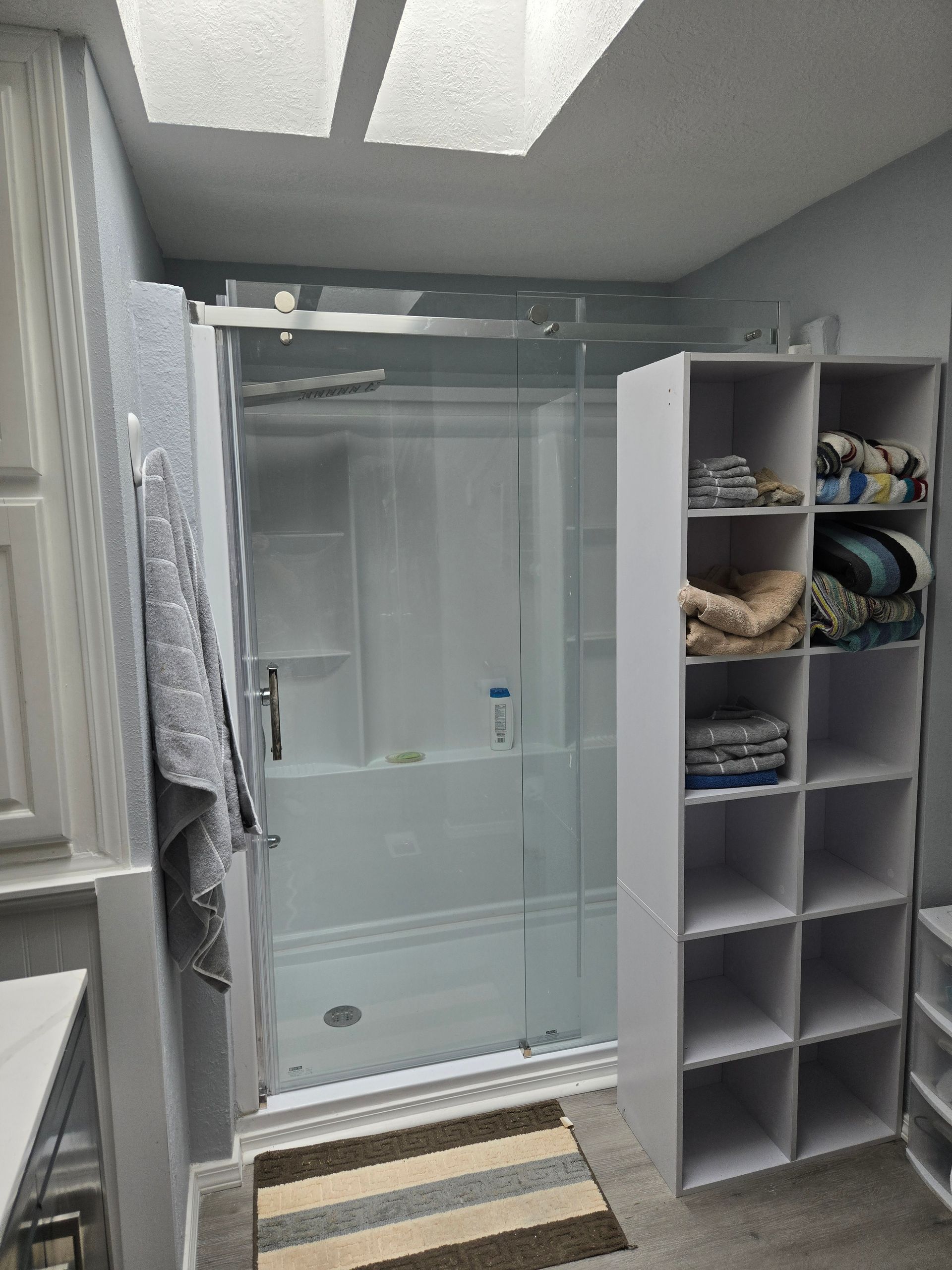 Bathroom with glass shower, gray walls, and a cubby shelf holding towels and toiletries.