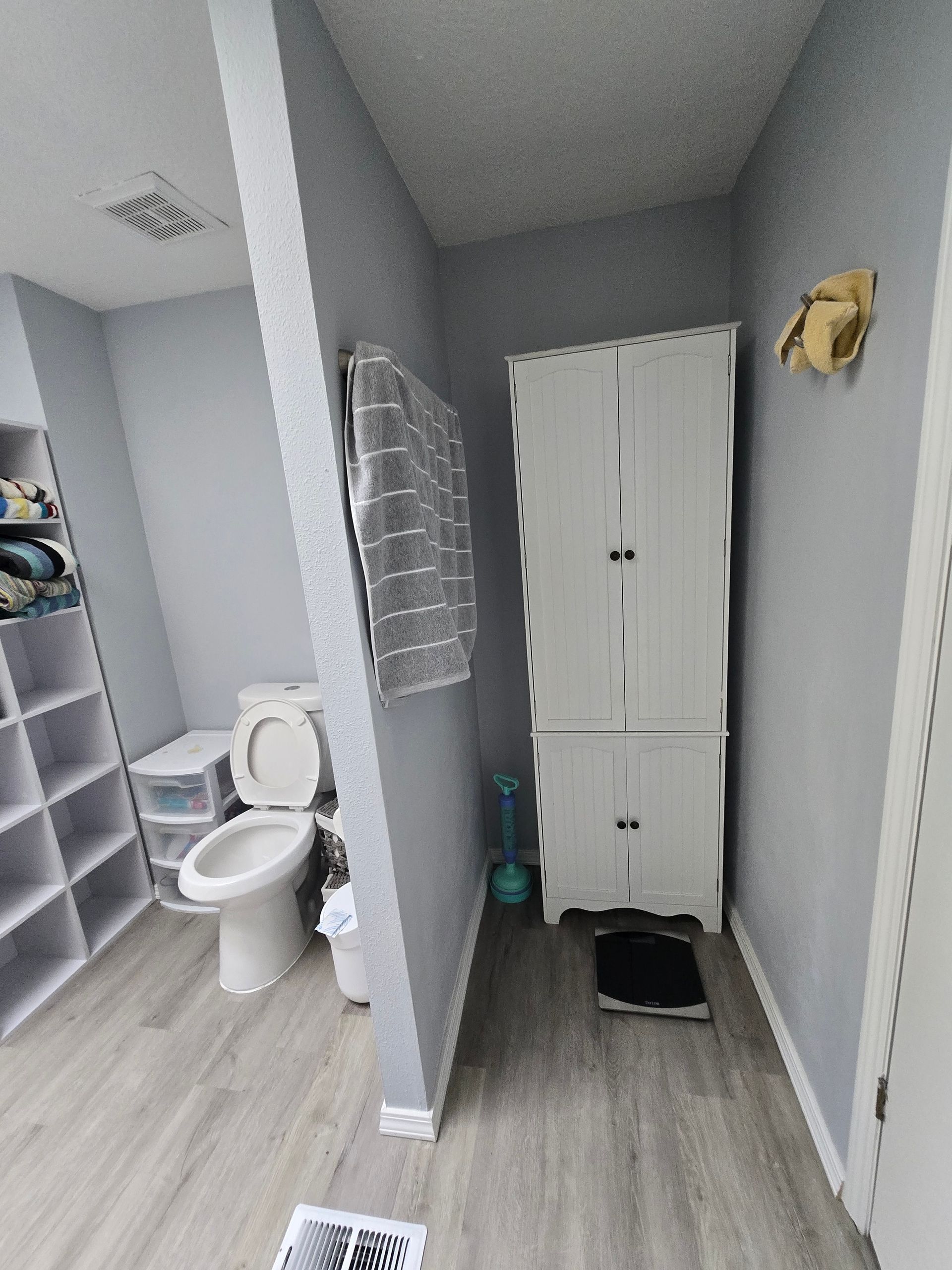 Small bathroom with open toilet, white cabinet, towel rack, and light wood floor.