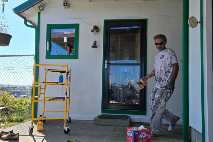 A person wearing paint-splattered clothes and sunglasses holds a power sander outside a white building with green trim.