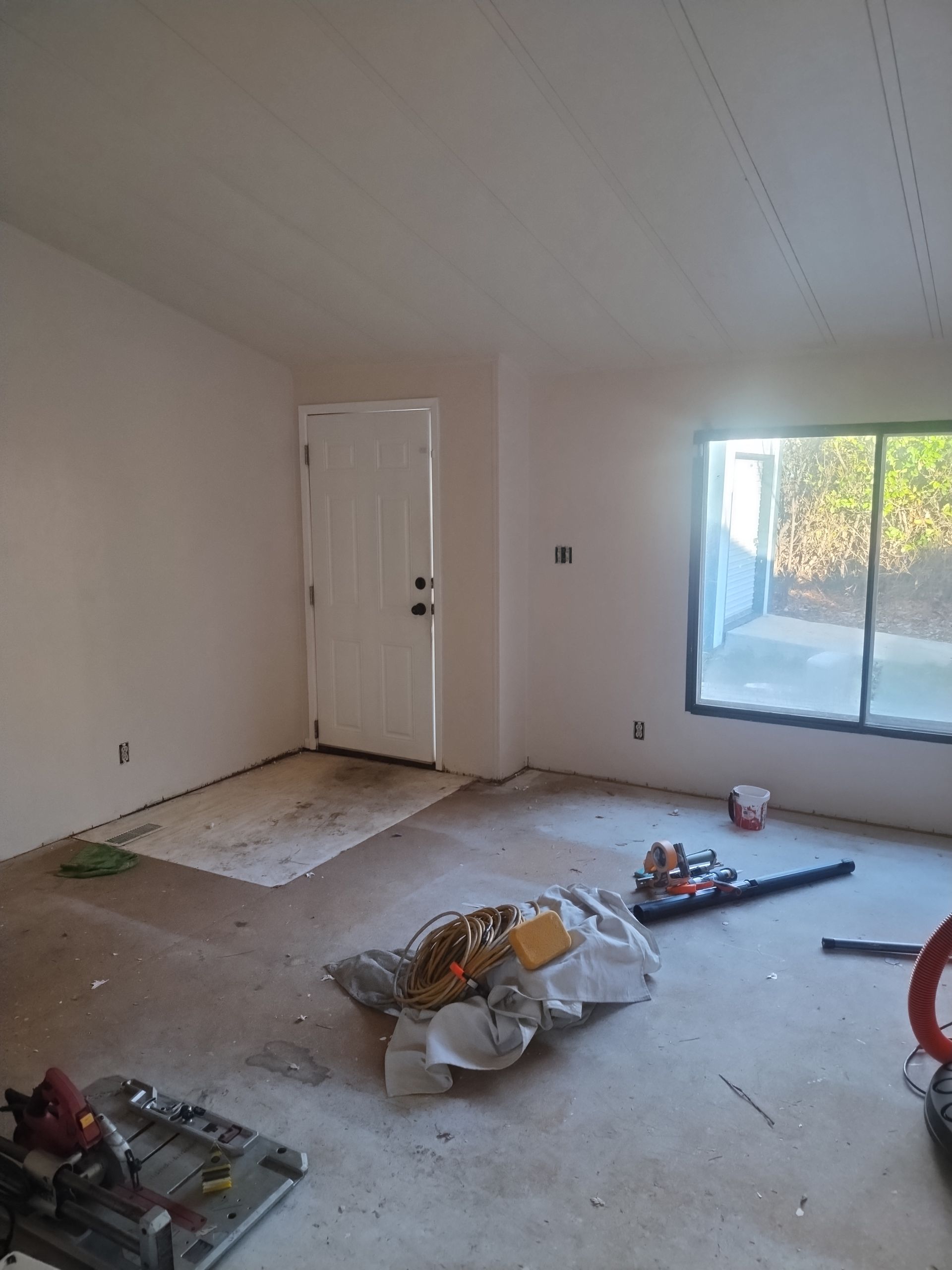 Empty unfinished room with bare floor, white walls, a closed door, and a window letting in daylight.