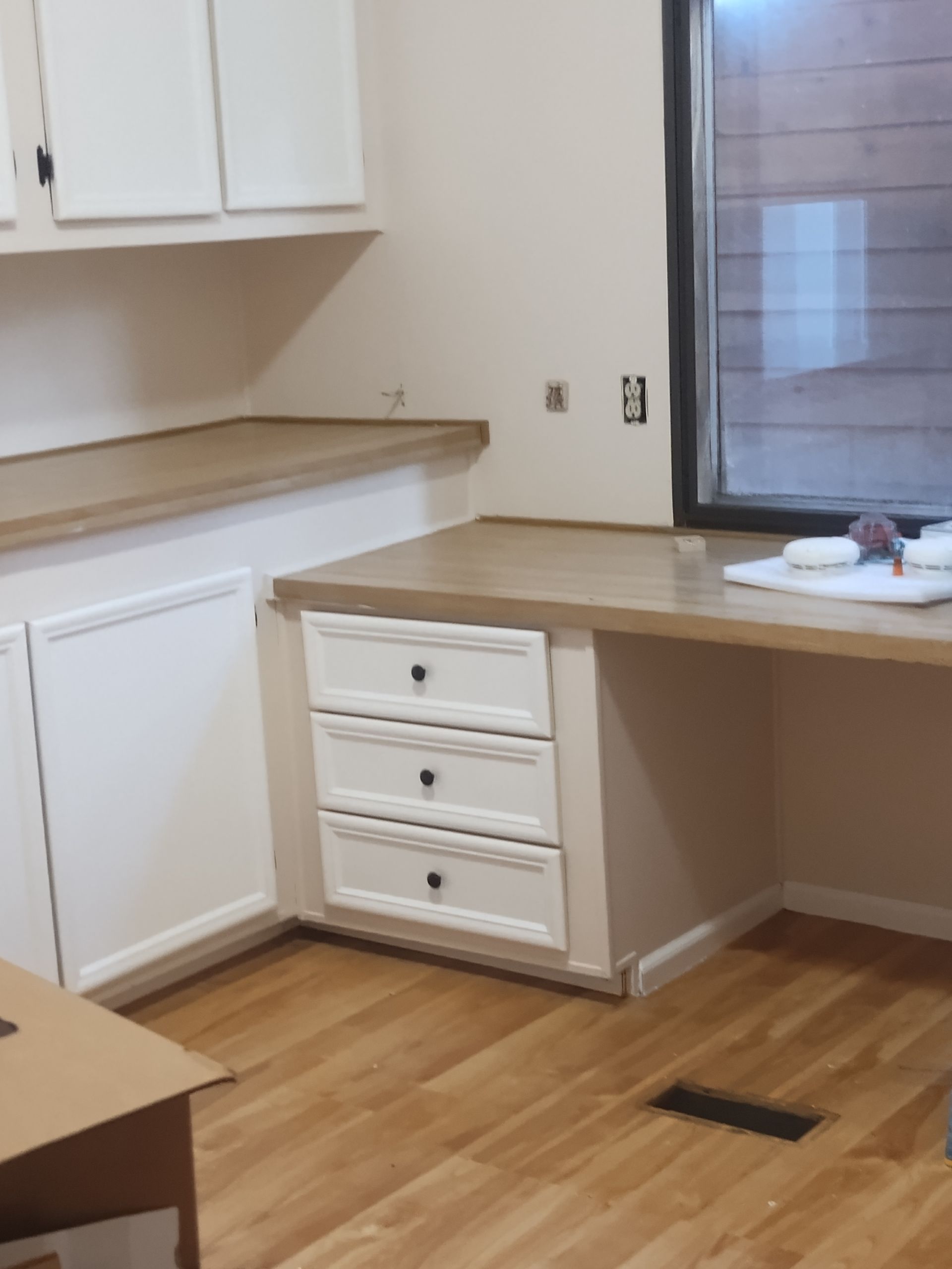Empty kitchen corner with white cabinets, light wood counters, and a window over the sink.