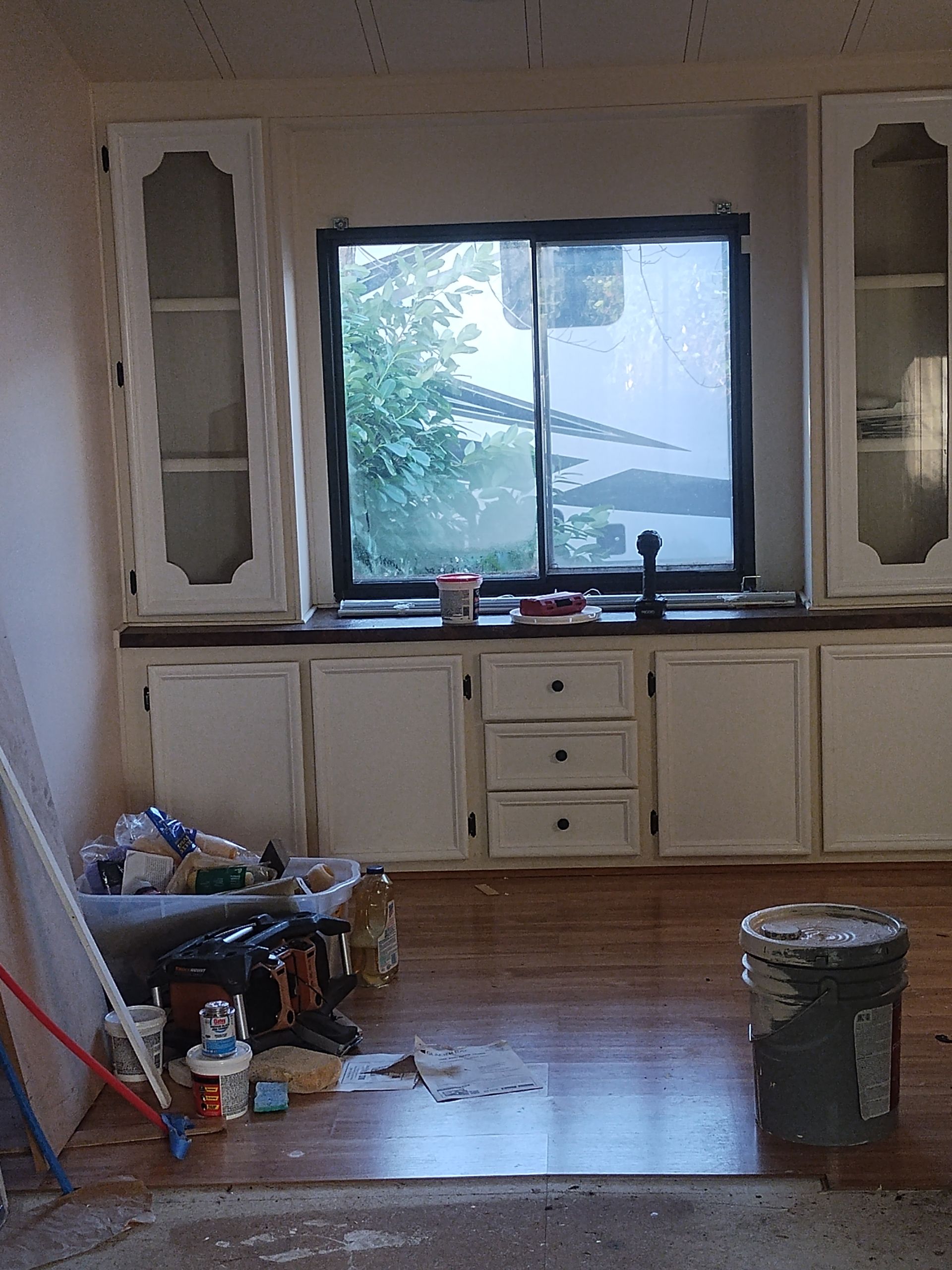 Empty kitchen with white cabinets, a large window, scattered items on the floor, and a bucket in front.