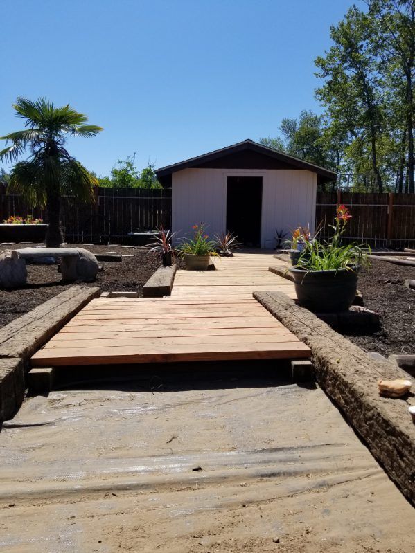 Backyard wooden deck leading to a small shed, with planters and rocks under a blue sky